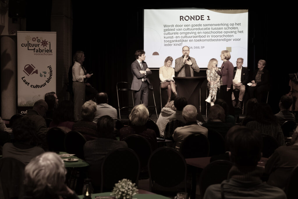 Een overzichtsfoto waarbij het publiek op de rug te zien is. Op het podium zijn drie politici in debat. Achter hen de overige politici gezeten op barkrukken. Daar weer achter de powerpoint slide met het onderwerp van de debatronde.