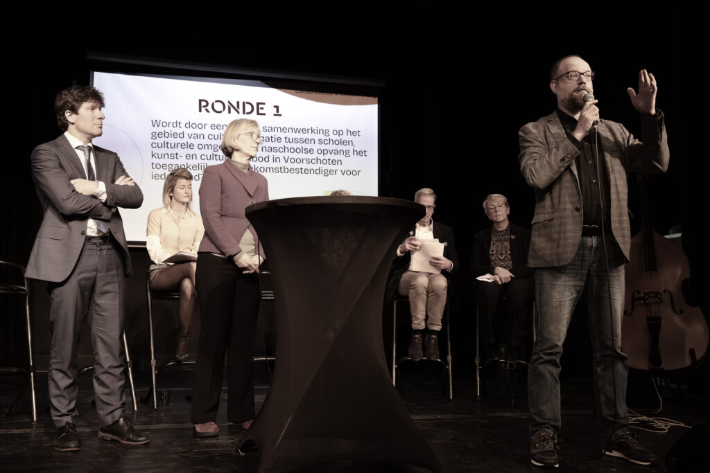 Foto genomen tijdens het Voorschotens Cultuurdebat. Op de voorgrond zijn drie politici met elkaar in debat. De overige politici zitten achter hen. Op de achtergrond is een powerpoint slide te zien waar het onderwerp van de ronde op staat.
